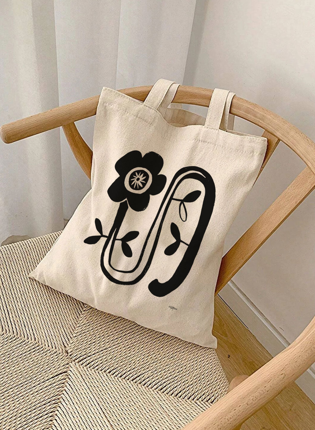 Tote bag with black floral design on a wooden chair against a white curtain background