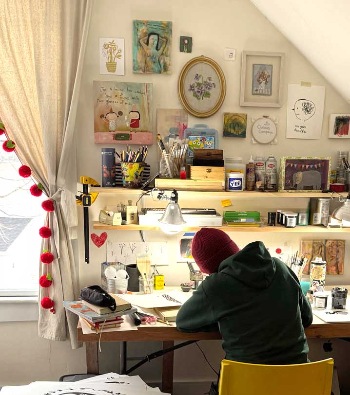 Artist sitting  in a cozy art studio with various items on shelves and a window with decorative curtains.
