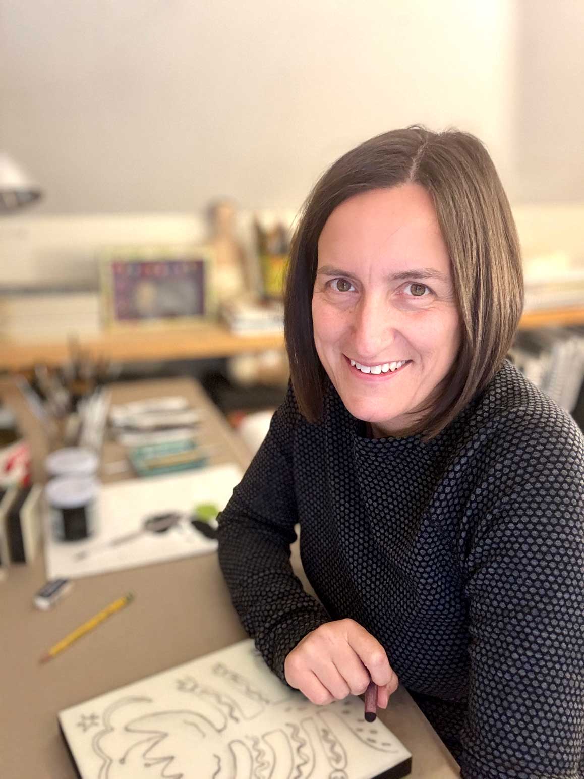 Person sitting at a desk with a drawing pad and pen, smiling.